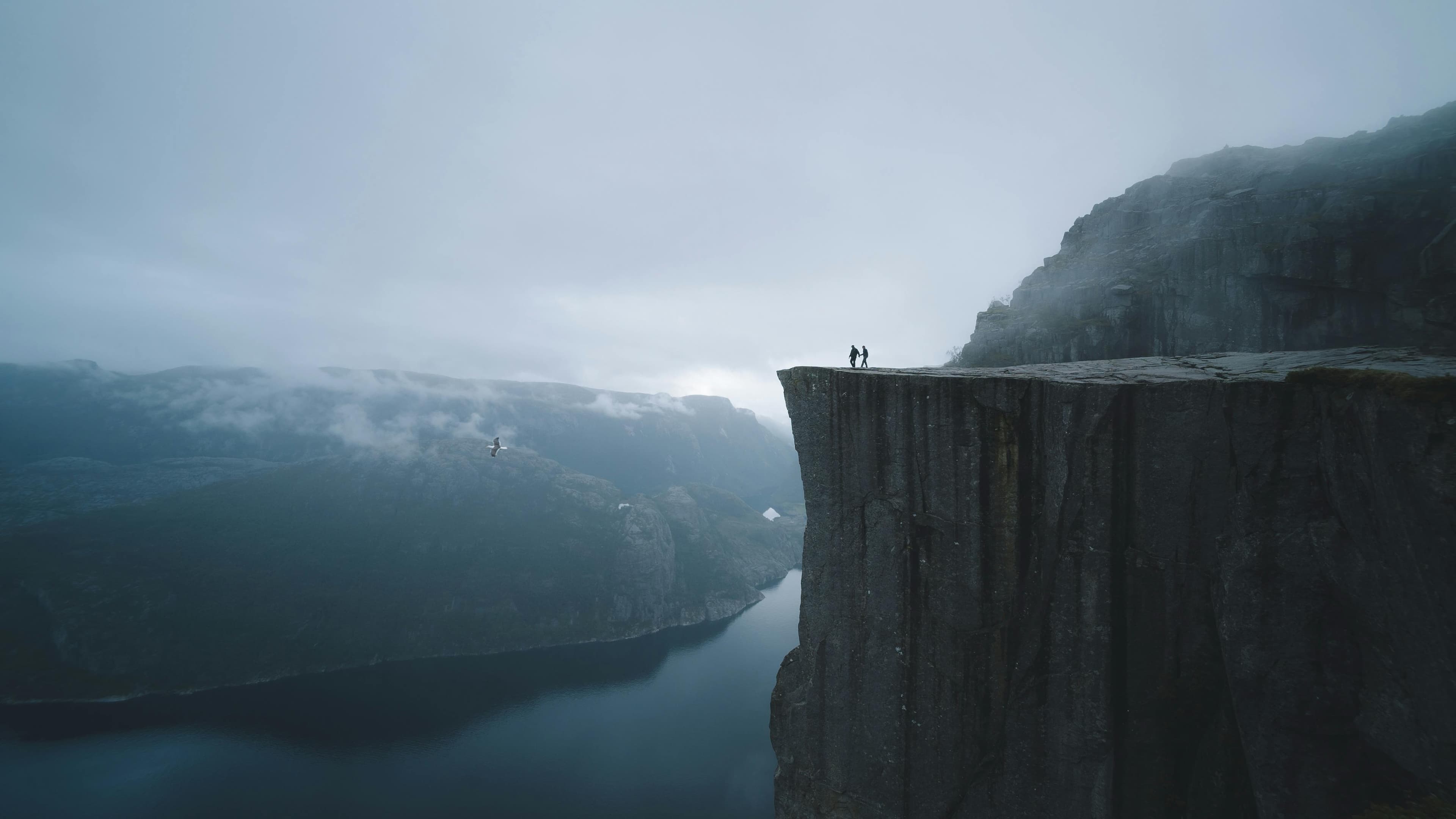 Two travellers standing on a dramatic cliff edge overlooking a fjord